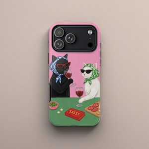 Puede incluir: Funda de teléfono rosa con una ilustración caprichosa de dos gatos con gafas de sol y pañuelos en la cabeza, sosteniendo copas de vino. Los gatos están sentados en una mesa verde con una caja roja que dice "SASSY".