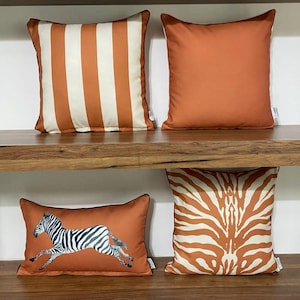 May include: Four decorative throw pillows on a wooden shelf. Two square pillows feature solid orange and orange and white stripes. The other two pillows have a zebra print and a running zebra illustration on an orange background.