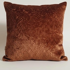 May include: A square, rust-colored decorative pillow with a textured, velvet-like surface. The pillow has a soft, plush appearance and is likely used for home decor. The color is a warm, earthy tone, suitable for various interior design styles.