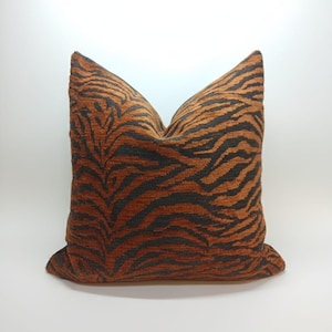May include: A square cushion with a tiger stripe pattern. The cushion features a brown and black design, with the stripes running vertically. The cushion is made of a soft, textured fabric and is a home decor item.