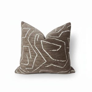 Op de afbeelding: Een vierkant decoratief kussen met een bruine, getextureerde achtergrond en een wit abstract lineair patroon. Het kussen heeft een neutrale kleur en zou verschillende woonstijlen aanvullen. Een zacht, comfortabel accentstuk.
