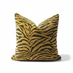 May include: Square decorative pillow with a tiger stripe pattern. The pillow features a mustard yellow background with black stripes. The pillow is made of a textured fabric and is designed for home decor.