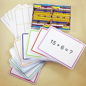 Puede incluir: Juego educativo de matemáticas con una caja de cuentas de colores y una pila de tarjetas didácticas. Las tarjetas didácticas tienen bordes de diferentes colores y problemas de matemáticas como "15 + 6 = ?" impresos. El juego incluye tarjetas en blanco con líneas.
