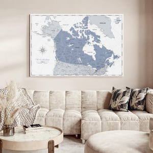 May include: A rectangular wall map print featuring a blue-toned Canada centered against a light gray background. The map includes detailed geographical labels, a compass rose, and the text The World Awaits in the lower left corner, all framed.