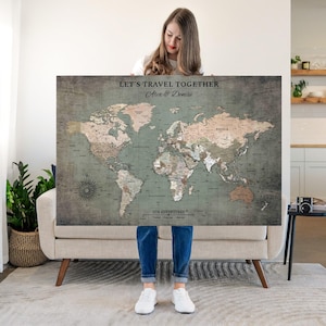May include: A large rectangular world map wall art piece with a muted sage green and beige color palette. The text "LET'S TRAVEL TOGETHER", "Alex & Denise", and "OUR ADVENTURES" is printed on the map. It features detailed continental outlines.