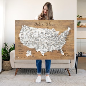 May include: A wooden map of the United States with white detailing, including state names and major cities. The map is labeled "Adventures of John & Maria" and includes text indicating travel plans. The map is held by a person.
