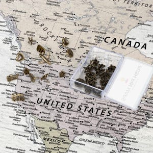 May include: A collection of bronze-toned metal push pins in various shapes including airplanes, hearts, stars, and houses, scattered across a printed map of North America alongside a clear plastic storage box labeled push pin maps.