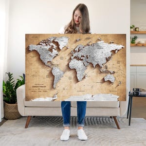 May include: A large rectangular world map wall art piece featuring a vintage-style tan background with raised, metallic silver-toned continents. The map includes printed text that reads The Travels of Gabriel & Denisse, along with various ocean and country labels.