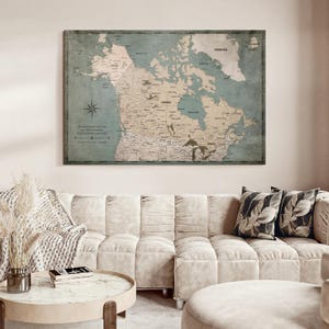 May include: A rectangular wall map of Canada and surrounding regions featuring a muted beige landmass against a dusty blue ocean background. The map includes detailed geographical labels, a compass rose, and printed text in the lower left corner.