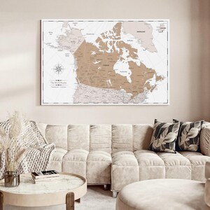May include: A rectangular wall map of the world featuring a prominent tan-colored Canada in the center. The map includes a compass rose, grid lines, and the text The World Awaits. The design uses a neutral color palette with white borders.