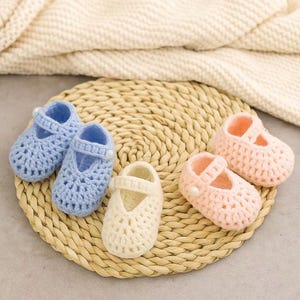 以下が含まれることがあります： 手作りのベビーシューズ。メアリージェーンスタイルの編み物で、水色、淡いピンク、クリーム色があります。靴は編まれたマットの上に置かれ、柔らかく質感のある背景があります。