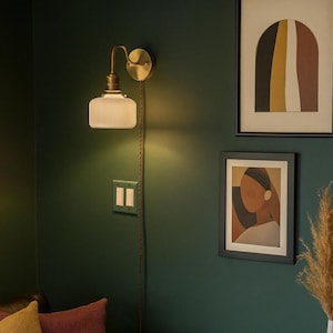 May include: A brass wall sconce with a ribbed glass shade casts light on a dark green wall. Two framed abstract artworks are above a black leather sofa with yellow and burgundy cushions. A white vase with dried plants sits on a black side table.