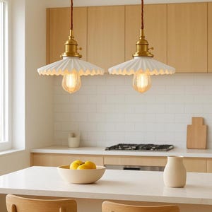 May include: Two pendant lights with white, scalloped shades and exposed Edison bulbs hang above a kitchen island. The lights have brass fixtures and are suspended by brown cords. The kitchen features light wood cabinets and a white countertop.