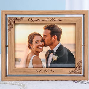 May include: A wooden picture frame with a photo of a couple kissing. The frame is light brown with decorative corner accents and the names "William & Amelia" and the date "6.8.2025" engraved on it. The photo is visible through a clear glass.