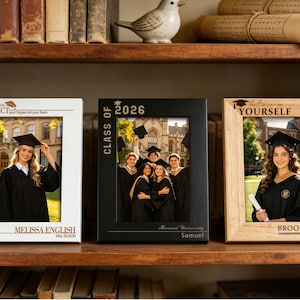 May include: Three graduation photo frames on a wooden shelf. The frames are white, black, and light brown. Each frame holds a photo of a graduate in a cap and gown. Text includes "REFLECT", "CLASS OF 2026", and "believe in yourself".
