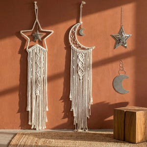 May include: Macrame wall hangings in star and crescent moon shapes, with a silver star and moon accents. The wall decor is cream-colored and hangs against a terracotta-colored wall. A wooden cube is in the foreground.