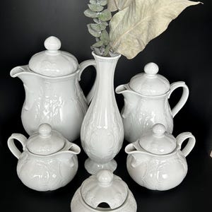 Può includere: Un servizio da tè in porcellana bianca con dettagli floreali ornati. Il set comprende due teiere, due piccole lattine, una zuccheriera e un vaso alto. I pezzi sono disposti su una superficie scura, con verde secco nel vaso.