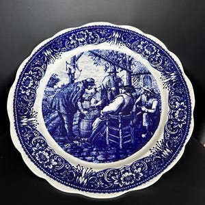 Könnte beinhalten: Ein runder, weiß-blauer Zierteller mit gewelltem Rand. Der Teller zeigt eine detaillierte blaue Illustration einer Gruppe von Menschen im Freien, mit einem floralen Bordürendesign.