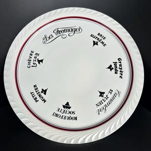 Può includere: Piatto da portata per formaggi in ceramica bianca con bordo rosso e bordo ondulato decorativo. Il piatto presenta il testo "Les Fromages" e nomi di formaggi come Brie Suprême e Camembert St. Julien.