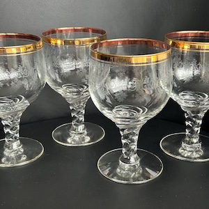 Op de afbeelding: Vier heldere wijnglazen met gouden randen en geëtste bloemmotieven. De glazen hebben gedraaide stelen en ronde bases, wat een elegante en klassieke uitstraling creëert. De gouden randen geven het glaswerk een vleugje luxe.
