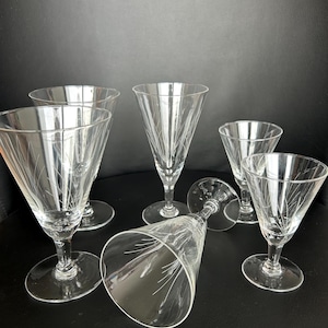 Könnte beinhalten: Ein Set aus sechs klaren Kristall-Weingläsern. Jedes Glas hat eine konische Schale mit geätzten linearen Designs und einen schlanken Stiel. Ein Glas liegt auf der Seite. Die Gläser stehen auf einer dunklen Oberfläche.