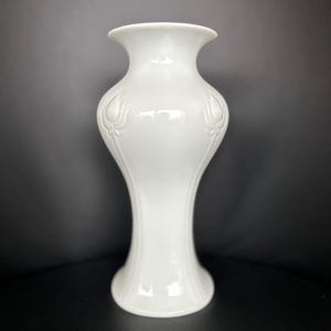 Könnte beinhalten: Eine weiße Keramikvase mit ausgestelltem Rand und breitem Boden. Die Vase hat eine glatte, glänzende Oberfläche und weist geprägte Blumendesigns an den Seiten auf. Die Vase ist etwa 25 cm hoch.