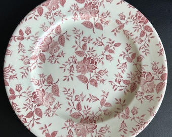 May include: A round, white ceramic plate with a repeating floral pattern in shades of red. The design features roses and leaves, creating a classic, vintage aesthetic. The plate is suitable for serving food.