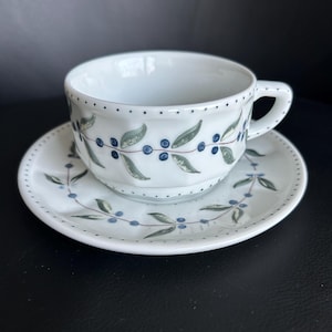 May include: White ceramic teacup and saucer set. The cup and saucer are decorated with a pattern of blue berries and green leaves. The rim of the cup and saucer are dotted with small blue dots. The cup has a handle.