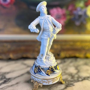 Könnte beinhalten: Eine weiße Porzellanfigur eines Mannes in Kleidung des 18. Jahrhunderts, die auf einem dekorativen Sockel steht. Der Sockel ist weiß mit goldenen und blauen Akzenten, getragen von verzierten goldenen Füßen. Die Figur ist etwa 15 cm hoch.