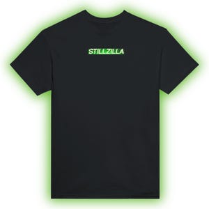 Könnte beinhalten: Schwarzes T-Shirt mit dem Text "STILL ZILLA" in Weiß und Grün. Der Text ist mittig auf der Brust platziert. Das Shirt hat kurze Ärmel und einen Rundhalsausschnitt.
