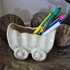 May include: A cream-coloured ceramic pen holder in the shape of a baby carriage, featuring two wheels and a scalloped top edge. The carriage contains several pens and markers in various colours, including yellow, green, blue, and pink. The text "C&B Operations LLC" is visible on one of the pens.
