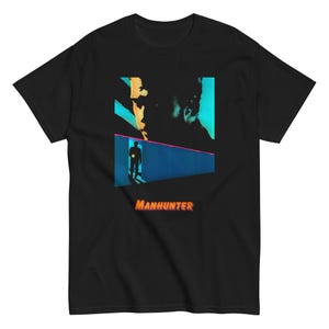 Puede incluir: Camiseta negra con un diseño gráfico que representa a una persona con una luz en un pasillo azul y turquesa. El diseño incluye una figura sombreada y la palabra "MANHUNTER" en naranja.
