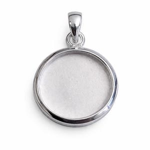 Puede incluir: Un colgante plateado con un marco circular y un lazo para una cadena. El colgante tiene un acabado pulido y un interior blanco en blanco, ideal para la personalización. La joya se muestra sobre un fondo blanco.