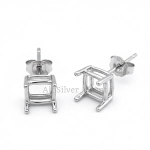 Może przedstawiać: Dwie srebrne kwadratowe kolczyki sztyfty. Każdy kolczyk ma czteronożną ramę ze sztyftem do włożenia. Kolczyki są umieszczone na białym tle, podkreślając ich minimalistyczny design i metaliczny połysk.