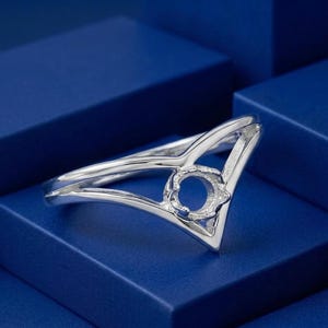 May include: A silver-toned metal ring featuring a V-shaped band design that splits into two parallel lines. At the center of the V-shape is a circular prong setting designed to hold a gemstone, showcasing a polished and reflective metallic finish.