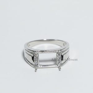 May include: A silver ring with a rectangular setting, designed to hold a gemstone. The ring features small, clear stones on either side of the setting. The words "All Silver Mount" are visible on the ring.