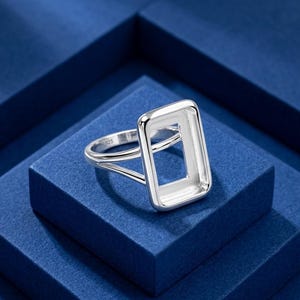 May include: A silver ring featuring a rectangular bezel setting with a hollow center. The band is split into two thin, polished silver segments that connect to the sides of the rectangular frame. The interior of the band is stamped with 925.