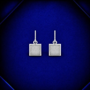 May include: A pair of silver-toned drop earrings featuring a square-shaped pendant with a textured, matte finish. Each earring is suspended from a simple hook wire, creating a clean and symmetrical design that hangs vertically from the top loop.
