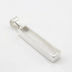 May include: A silver-toned rectangular pendant featuring a long, narrow, recessed channel with a textured, matte-finish base. The pendant includes a polished, smooth bail at the top for attachment to a chain, showcasing a clean, metallic design.