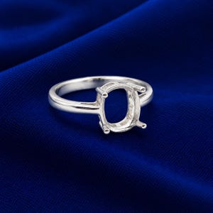 Peut inclure: Une bague en argent poli dotée d'un chaton ovale vide à quatre griffes conçu pour accueillir une pierre précieuse. L'anneau est lisse et arrondi, présentant une finition métallique qui reflète la lumière sur toute sa surface.