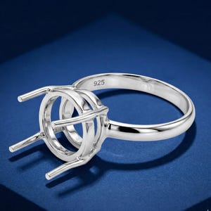 May include: A silver ring setting with a round center and four prongs to hold a gemstone. The ring is marked with "925" and is set against a blue background. The ring is made of a shiny, polished metal.