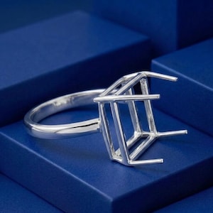 May include: A polished silver ring setting featuring a rectangular four-prong head designed to hold a gemstone. The metal band is smooth and circular, connecting to a structured, open-frame basket mount that extends upwards to secure a stone.