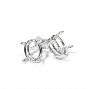 May include: A pair of silver-colored earring settings with an oval-shaped bezel. The settings have four prongs and a post for securing to the ear. The earrings are displayed on a white background.