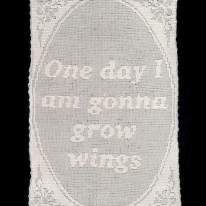 May include: Cream-coloured, rectangular crochet piece featuring the text "One day I am gonna grow wings". The text is within an oval border, with floral designs at the corners. The background is black.