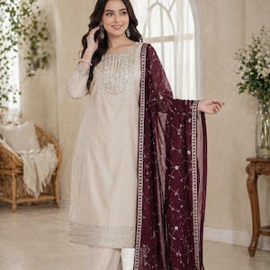 Puede incluir: Conjunto salwar kameez color crema con intrincados bordados en el escote y el dobladillo. Un dupatta burdeos oscuro con motivos florales y un borde plateado complementa el atuendo. Atuendo tradicional indio para ocasiones festivas.