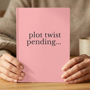 May include: A pink hardcover notebook with the text "plot twist pending..." in a dark gray font. The notebook is held open by two hands on a wooden surface.