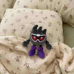 Puede incluir: Un peluche con forma de criatura gris oscuro con ojos rojos y botas moradas. El juguete tiene un asa blanca y está sobre un fondo con estampado floral.