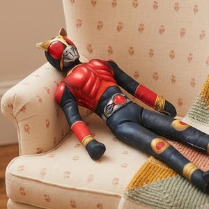 May include: A plush toy of a character in a red and black costume with a silver mask and gold accents. The toy is lying on a floral patterned chair with a colorful blanket.