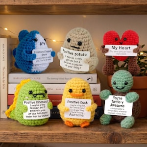 May include: A collection of crocheted amigurumi figures, including a blue shark, a potato, a red heart, a green dinosaur, a yellow duck, and a green turtle. Each figure holds a small sign with positive messages.
