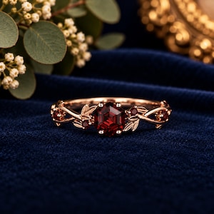 Op de afbeelding: Een roségouden ring met een centrale zeshoekige rode granaat edelsteen, geflankeerd door kleinere ronde rode granaten. De ringband is ontworpen met gedetailleerde metalen blad- en rankmotieven die vanaf de zetting langs de band lopen.